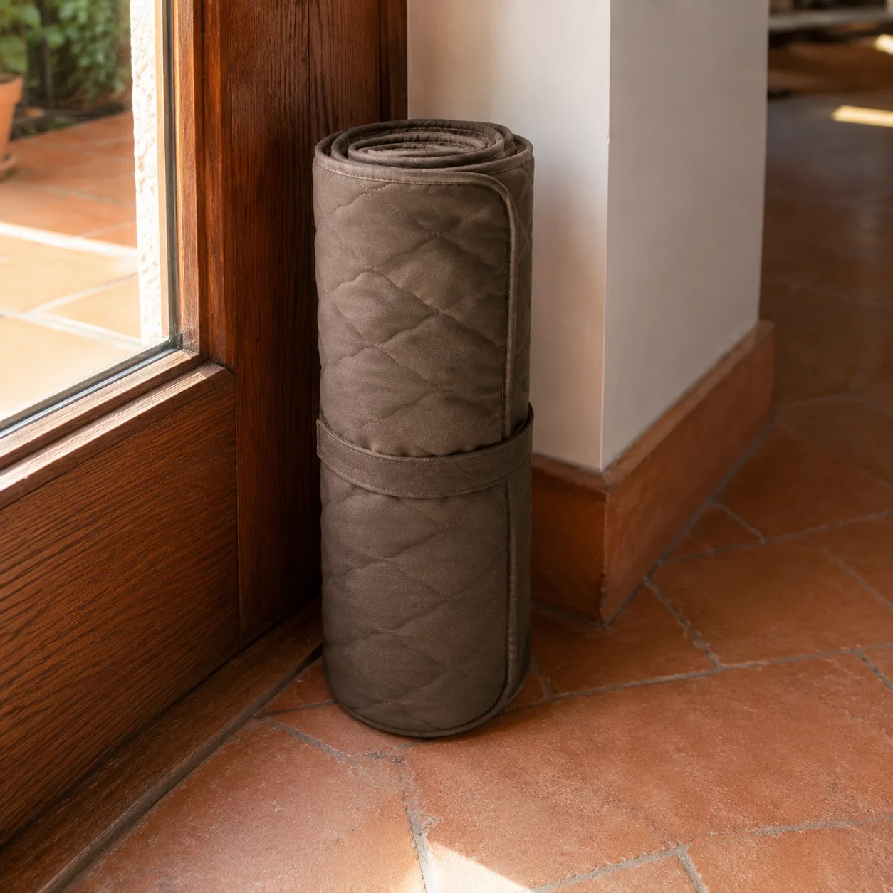 A brown Frimouf Ice cooling mat rolled up and strapped, standing by a glazed door on a terracotta tile floor